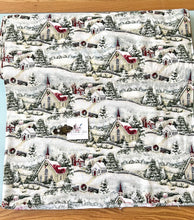 Load image into Gallery viewer, Susan Wi get Country Church Christmas fabric with a repeating Christmas village pattern featuring snow-covered houses, trees, and fences on a white background, laid flat on a wooden surface.