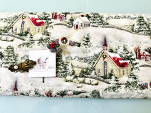 Load image into Gallery viewer, A close-up of a Susan Winget Country Church Christmas fabric featuring a Christmas village print with snow-covered hills, churches, trees, and houses in red, green, cream tones. A small white card and decorative embellishments are placed on top of the fabric.