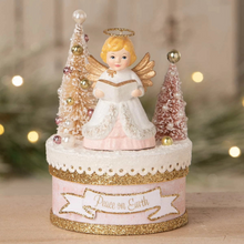 Load image into Gallery viewer, A decorative angel figurine on a round base, reading a book, with two small frosted Christmas trees and a halo, set against a softly blurred holiday background with warm lights.