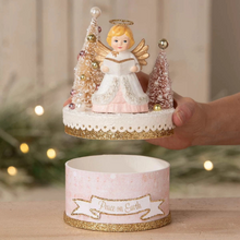 Load image into Gallery viewer, A hand holds a round Christmas ornament or decorative piece featuring a small angel figurine with gold wings and halo, flanked by two glittery Christmas trees, above a matching cylindrical container labeled 'Peace on Earth.' The background is softly blurred with warm lights and greenery.