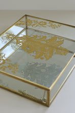 Load image into Gallery viewer, A rectangular glass jewelry box with a gold metal frame and gold floral motifs printed on the mirrored lid, shown on a light neutral background.