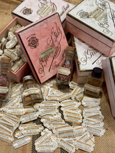 Load image into Gallery viewer, A collection of vintage-style perfume and fragrance bottles with many matching paper labels scattered in front, alongside decorative boxes and cartons printed with elegant script and crest designs.