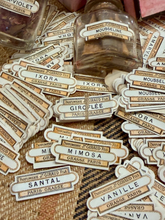 Load image into Gallery viewer, A collection of vintage-style perfume label stickers scattered on a woven brown fabric background, with a small clear glass bottle containing amber liquid placed near the top center.