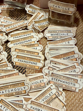 Load image into Gallery viewer, A pile of vintage-style perfume label stickers with French names like Vanille, Frangipane, Maréchale, and Violet, printed on cream and tan paper with black text, arranged on a woven beige surface.