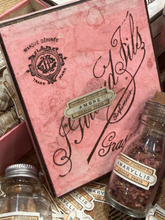 Load image into Gallery viewer, A vintage-style pink decorative box or package with black script text reading 'Madame Tails' and a small label that says 'Ambré Grand Paris,' surrounded by small glass bottles and scattered matching labels on a textured surface.