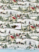 Load image into Gallery viewer, A close-up of a Susan Winget Country Church Christmas-themed fabric print featuring snow-covered hills, small houses, churches, trees, and wreaths in white, red, green, and beige tones. A small white card with a logo is placed over the center of the fabric.