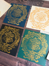 Load image into Gallery viewer, Four square decorative cards or prints arranged on a dark wood surface, each showing an ornate gold damask-style floral pattern over a different background color: blue, cream, black, and teal green.