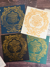 Load image into Gallery viewer, Four square decorative cards or prints arranged on a dark wood surface, each featuring an ornate gold damask-style floral motif over a different base color: navy blue, off-white, black, and teal green.
