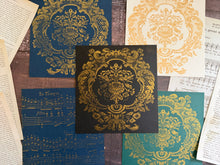 Load image into Gallery viewer, A flat-lay arrangement of decorative printed cards or paper sheets with ornate damask-style motifs in gold, blue, teal, and cream, placed on a rustic wood surface with scattered vintage book pages around them.
