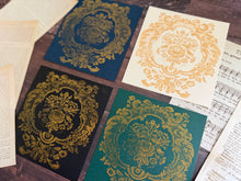 Load image into Gallery viewer, Four square art prints with ornate gold damask floral designs on different colored backgrounds, arranged on a dark wood surface with vintage book pages and sheet music.