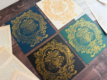 Load image into Gallery viewer, Four square decorative cards or paper pieces arranged on a dark wood surface, each featuring an ornate gold damask-style floral pattern on a different background color: navy blue, cream, teal green, and black. Fanned book pages surround the pieces.