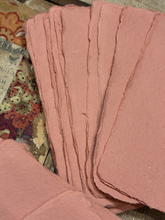 Load image into Gallery viewer, A stack of handmade, unbleached pink paper sheets fanned out, showing their rough, rustic edges, resting on a patterned fabric surface.