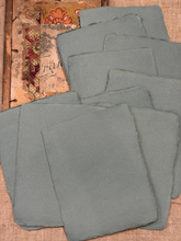 Load image into Gallery viewer, A stack of slate coasters with rough, natural edges, displayed on a textured burlap surface next to a vintage patterned book.