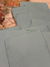 Load image into Gallery viewer, A stack of rectangular gray felt or fabric squares laid out on a patterned surface with a beige and orange decorative paper visible in the upper left corner.