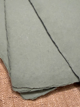 Load image into Gallery viewer, A close-up of overlapping handmade paper sheets with irregular rag edges, resting on a woven tan fabric surface.