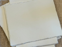 Load image into Gallery viewer, A stack of unbleached, handmade paper sheets with textured, slightly uneven edges, displayed on a brown woven fabric background.