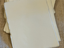 Load image into Gallery viewer, A stack of blank, off-white handmade paper sheets with rough, unbleached edges, resting on a textured beige fabric surface.