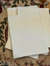 Load image into Gallery viewer, A stack of blank white paper sheets with rough, handmade edges, resting on a patterned fabric surface.
