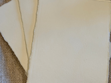 Load image into Gallery viewer, Three sheets of handmade cream-colored paper with rough, torn edges, fanned out over a textured brown fabric background.