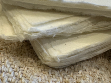 Load image into Gallery viewer, A close-up of a stack of off-white, textured felt or fleece material resting on a beige woven rug.