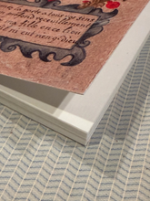 Load image into Gallery viewer, A close-up of a printed page or card with decorative black script and red floral accents on a warm pinkish-brown background, resting on a blue-and-white striped textured surface.