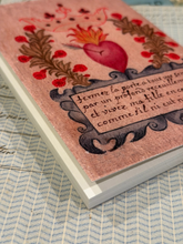 Load image into Gallery viewer, A close-up of a decorative book or notebook cover featuring a pink background, a red heart with flames, dark brown floral garlands, and a black ornamental cartouche with handwritten French text. The item is photographed at an angle on a blue-and-white patterned surface.