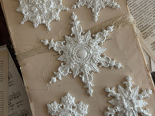 Load image into Gallery viewer, White ornate snowflake embellishments arranged on a vintage beige book or album page with printed sheet music visible around the edges.