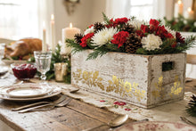 Load image into Gallery viewer, A rustic white wooden box centerpiece decorated with gold leaf patterns, filled with a holiday floral arrangement of red and white flowers, greenery, and pinecones, set on a decorated dining table with candles and plates.