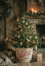 Load image into Gallery viewer, A decorated Christmas tree in a vintage Santa-themed bucket, styled in a cozy rustic living room with a stone fireplace, candles, wrapped gifts, and pinecones.