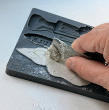 Load image into Gallery viewer, A close-up of a hand using a metal spatula to scrape light gray plaster or clay into a black mold tray, with embossed text including 'TOD Belsmoke' and 'JUEU' visible on the tray.