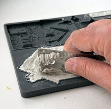 Load image into Gallery viewer, A close-up of a hand using a metal ruler to measure a layer of gray plaster or clay on a black plastic 3D printing mold tray.