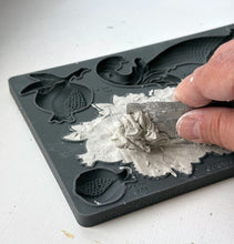 Load image into Gallery viewer, A close-up of a hand using a small metal scraper to remove wet gray plaster or clay from a dark gray silicone mold tray with fruit-shaped cavities.