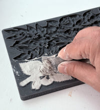 Load image into Gallery viewer, A close-up of a hand pressing a gray stamping ink pad into a black floral rubber mold, with the embossed design becoming visible on the surface.