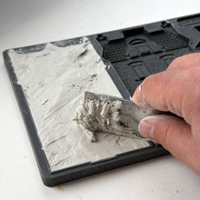 Load image into Gallery viewer, A close-up of a hand using a metal ruler to measure a layer of gray plaster or clay on a black square tray, with the textured material spread across one half of the tray.