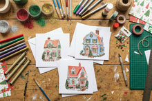 Load image into Gallery viewer, Three small illustrated Christmas house cards are arranged on a wooden craft table surrounded by art supplies, including paintbrushes, glitter, tape, markers, and a green cutting mat.