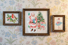 Load image into Gallery viewer, Three framed vintage-style needlepoint artworks hanging on a floral wallpaper background. The center, largest framed piece shows Santa Claus beside a Christmas tree and presents; the left smaller frame shows a bird with holly and roses; the right smaller frame shows a ballerina.