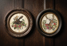 Load image into Gallery viewer, Two round framed wall art pieces hanging on a dark wood paneled wall. The left frame shows a moose surrounded by flowers, and the right frame shows a deer surrounded by flowers, both in ornate dark wood frames.