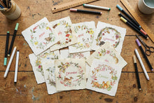 Load image into Gallery viewer, A flat-lay arrangement of handmade paper cards featuring monthly winter animal illustrations and names, displayed on a rustic wooden table surrounded by art supplies like markers, scissors, and rulers.