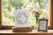 Load image into Gallery viewer, A white art print card featuring a floral wreath and the word 'June' in the center, displayed on a rustic wooden block on a windowsill. Next to it is a small ornate silver picture frame and a glass jar filled with colorful wildflowers, all set against a soft, blurred green garden background.