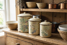 Load image into Gallery viewer, Three vintage cream enamel canisters with lid lids decorated with botanical and bird illustrations, displayed on a rustic wooden kitchen cabinet alongside matching bowls and copper pots.