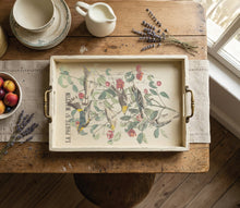 Load image into Gallery viewer, A rectangular serving tray with brass handles, featuring a vintage-style botanical illustration of birds and cherries with the text 'LA PORTS S. MARTIN', displayed on a rustic wooden table with a placemat, fruit bowl, lavender, and cream-colored ceramic items.