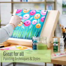 Load image into Gallery viewer, Person painting a colorful floral scene on an easel with art supplies around.