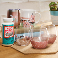 Load image into Gallery viewer, Two glitter-dipped wine glasses on a wooden surface with Mod Podge Dishwasher Safe Glue Sealer Finish bottle and floral mug in the background.