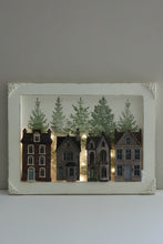 Load image into Gallery viewer, A square decorative shadow box or framed wall art piece featuring four miniature house cutouts in front of green pine trees, with warm string lights glowing behind them, set against a plain light gray wall.