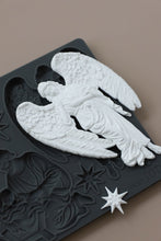 Load image into Gallery viewer, A white angel figurine with detailed wings and draped robes displayed on top of a dark gray decorative plaque featuring embossed holiday motifs and a small star piece, set against a solid taupe background.
